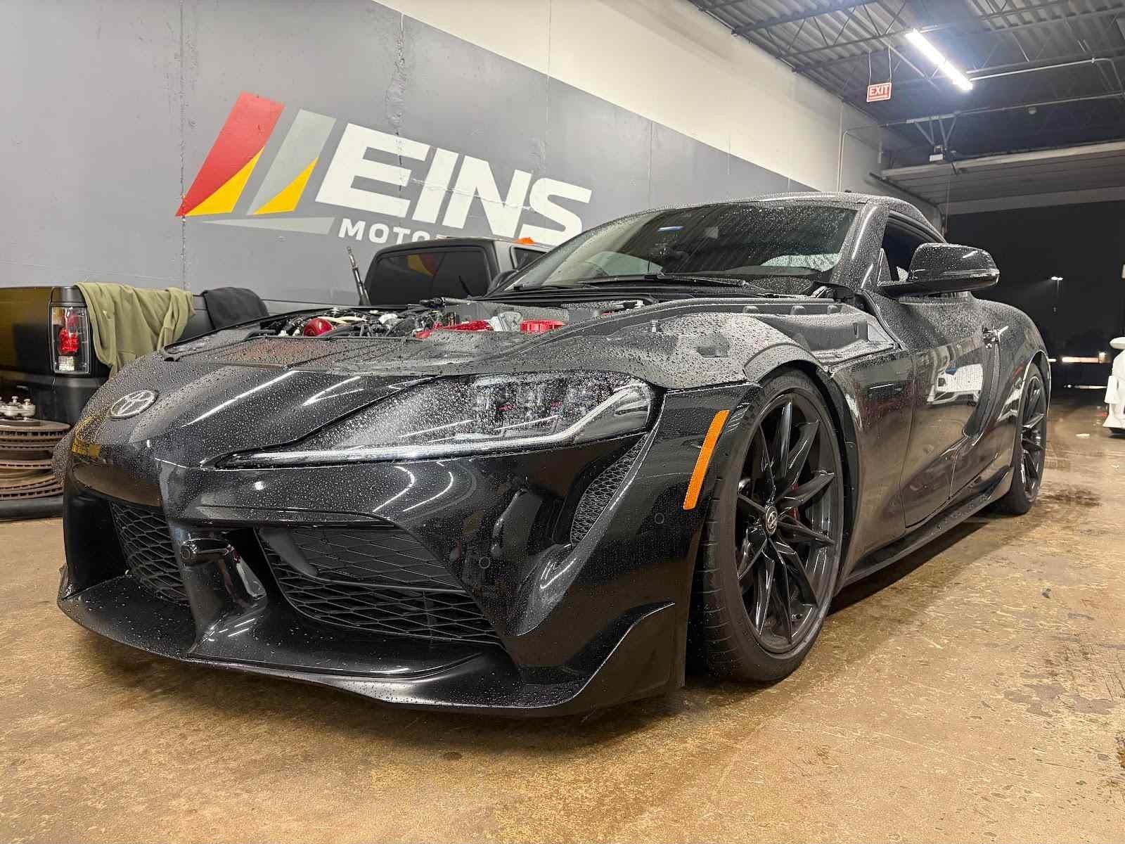 Black GR Supra, hood off, in front of the Eins wall